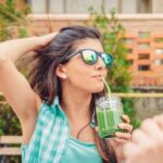 Beautiful young woman with sunglasses drinking green vegetable smoothie with straw in a summer day outdoors. Healthy organic drinks concept.